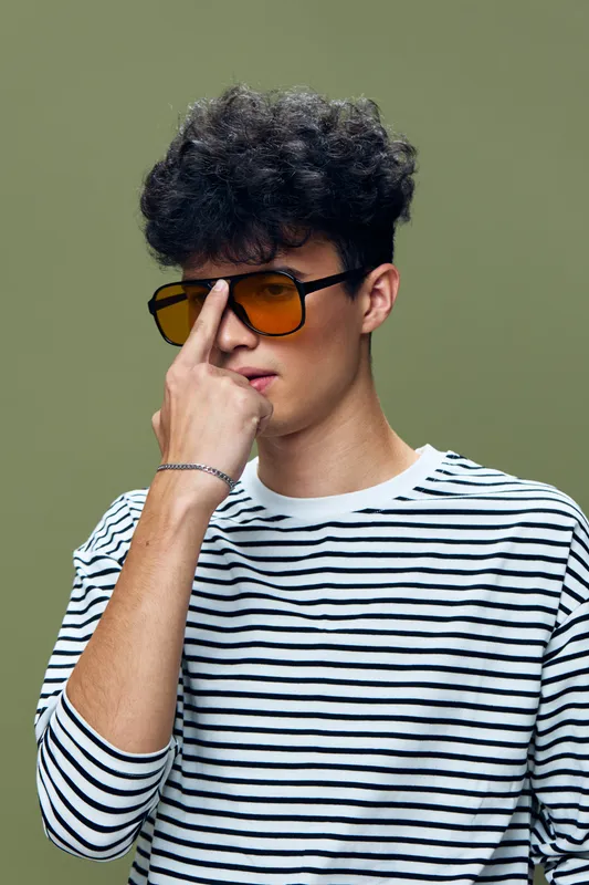 Young man with curly hair wearing orange sunglasses and striped shirt, posing confidently against a muted green background in contemporary casual style.