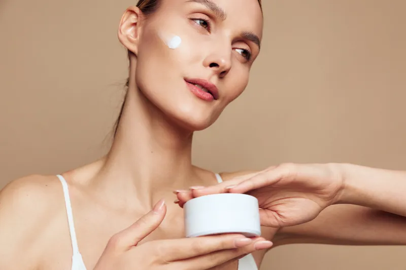 Young woman applies moisturizing cream on face holds white jar skincare product looks relaxed with clear skin isolated on beige background natural beauty concept closeup portrait.