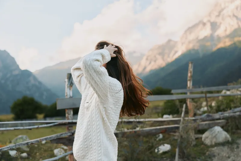 A young woman in a white knit sweater stands outdoors among mountains, hair blown by wind, facing away as she enjoys the dramatic landscape.