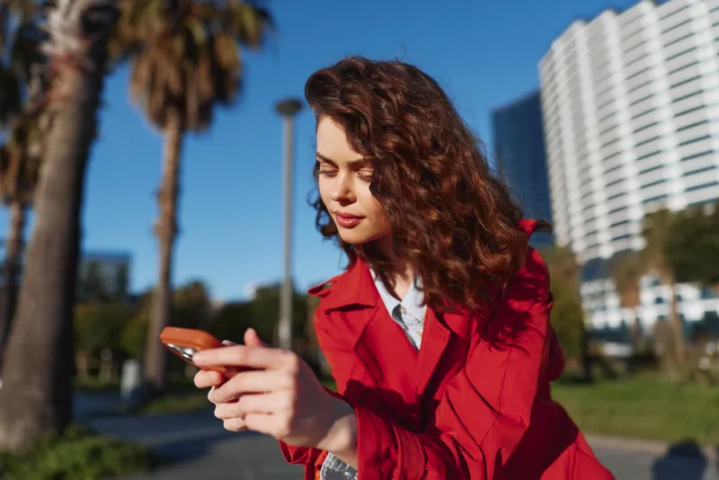 Young woman with curly hair wearing a red coat looks at her smartphone outdoors on a sunny day with palm trees and modern buildings in the background.