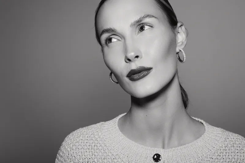 Portrait of a young white woman with light skin and slicked-back hair wearing hoop earrings and a textured sweater. The black and white photo highlights elegance and fashion concepts.