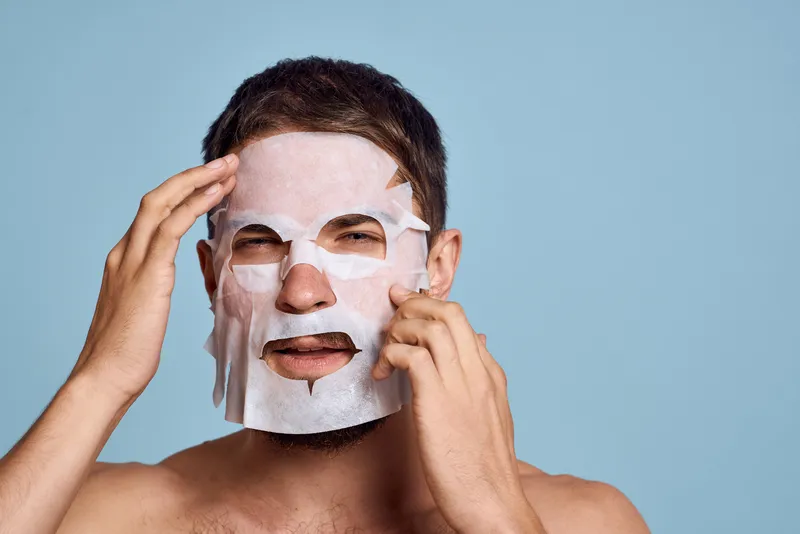 Мужчина в очищающей маске на голубом фоне жестикулирует руками, обнаженный торс, обрезанный вид