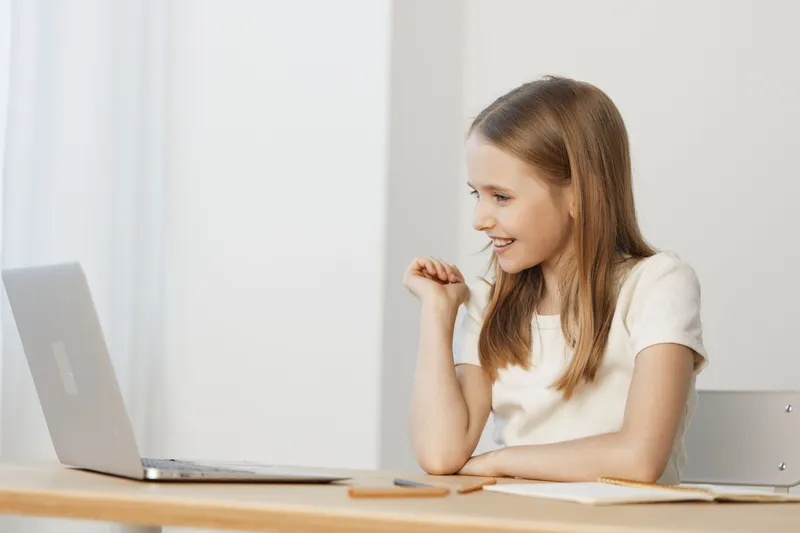 Smiling young girl engaging with a laptop in a bright, minimalist workspace, embodying joy and curiosity while working on school projects or online learning