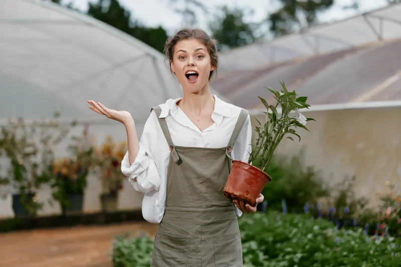 Joyful young woman in a garden holding a plant, wearing casual overalls and a white shirt, with vibrant greenery in the background, representing gardening, growth, and nature