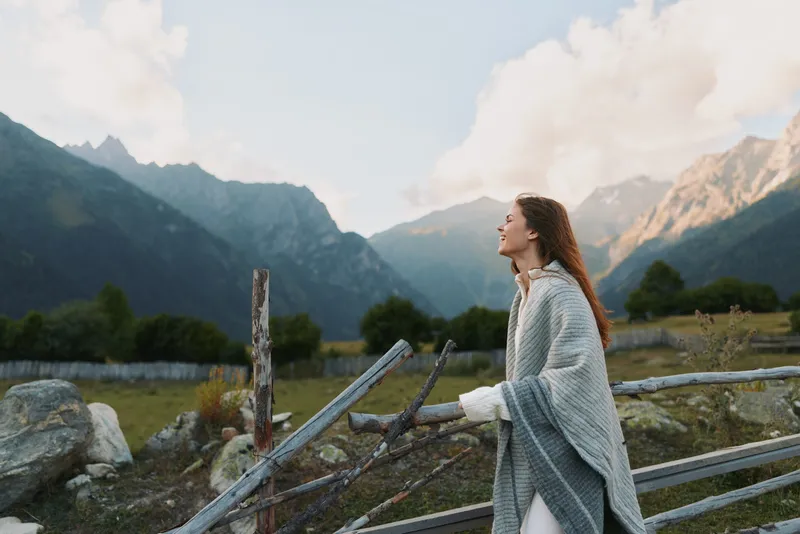 A woman wrapped in a cozy blanket stands outdoors near a wooden fence with mountains in the background, enjoying a peaceful moment in nature lifestyle.