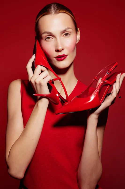 Young woman in red sleeveless top holds red high heel shoe near face on red background. Concept of fashion, style, elegance, and bold color contrast in studio lighting.