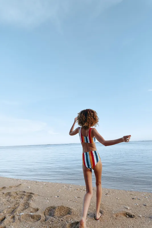 A young girl in a striped swimsuit stands with her back to the camera on a sandy beach, facing calm water under a clear blue sky, embodying carefree summer mood.
