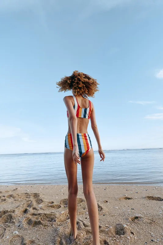Young woman with curly hair in striped swimsuit stands on sandy beach facing calm ocean horizon, bright daytime sky, relaxed mood, travel and summer lifestyle concept, vacation alone