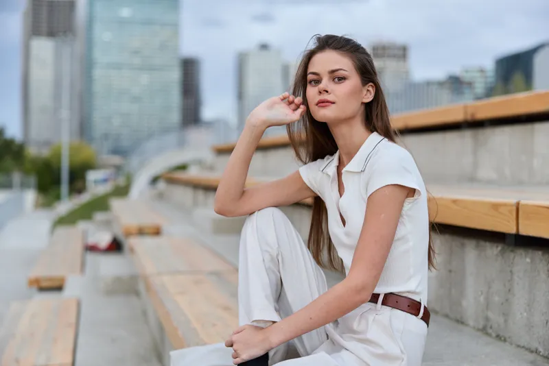 Confident businesswoman sitting outdoors, wearing a stylish white outfit and conveying professionalism and poise in an urban setting.