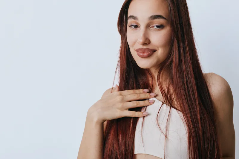 Young woman with long red hair and light skin smiling gently with hand on chest against plain background. Beauty, emotion, and natural expression in portrait style.