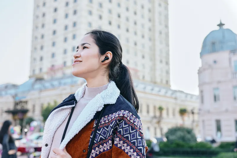 Woman with headphones gazing upward in the city, wearing a patterned jacket and scarf, urban street scene with modern architecture and bright daylight, her smile conveys confidence, travel vibe and