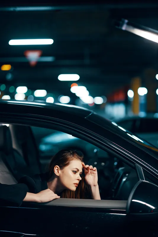Вертикальная фотография, снятая сбоку ночью: женщина сидит в чёрном автомобиле и смотрит в окно, глядя в боковое зеркало, при этом держит руку возле лица