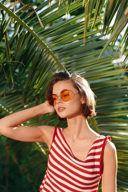A stylish woman stands outdoors among lush tropical palm leaves, wearing a red and white striped sleeveless top and round sunglasses. She exudes confidence and a sunny, carefree vibe.
