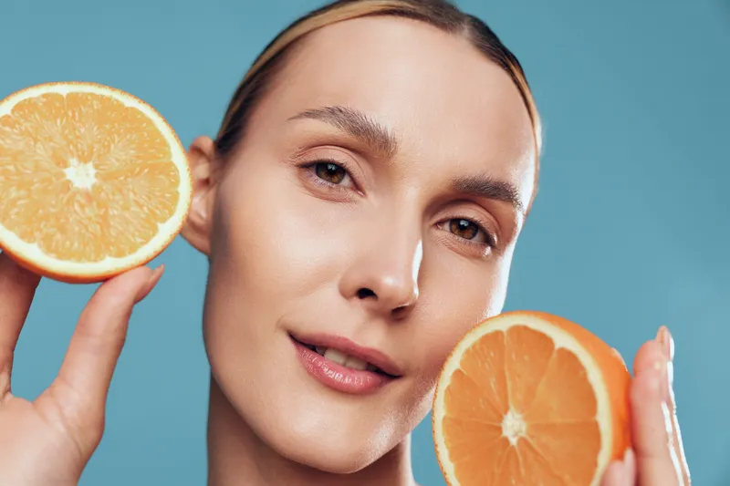 Young woman with light skin holds orange halves near her face, highlighting citrus inspired skincare and a fresh wellness vibe in a bright studio against a blue background.