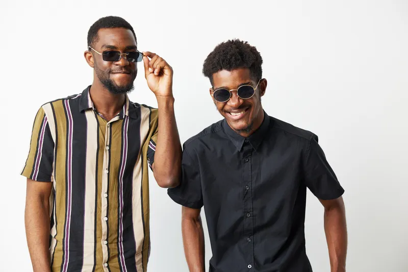 Two African American men wearing stylish sunglasses pose with cheerful expressions, showcasing modern fashion in a minimalist setting.