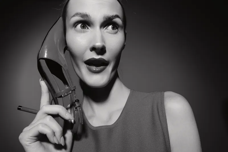 Black and white portrait of a pale white woman holding a high heel shoe near her ear as if it is a phone, with surprised expression on her face in studio lighting.