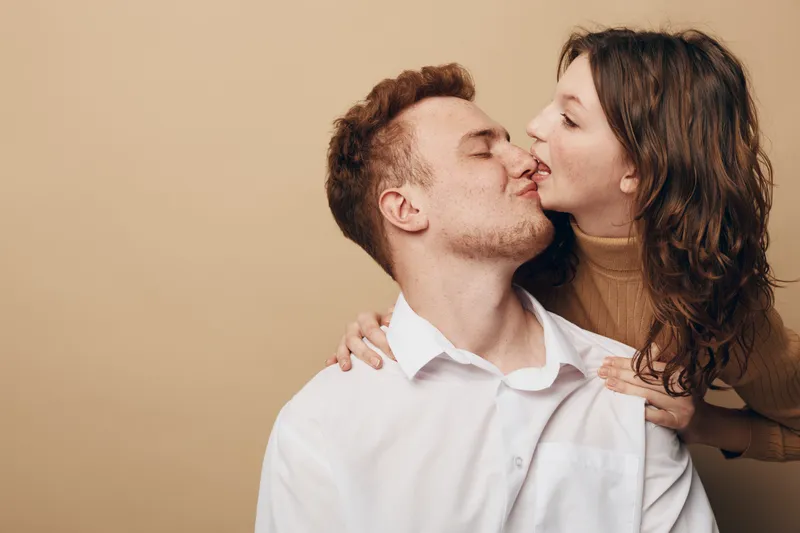 Young couple in casual clothes shares intimate moment, woman gently kisses man on cheek. Warm beige background creates cozy romantic atmosphere and connection between partners.