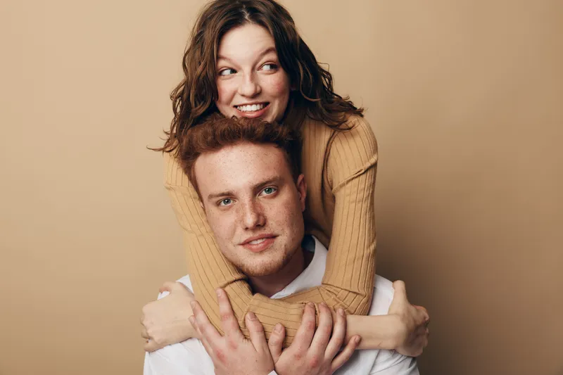 Smiling young couple with woman hugging man from behind on beige background. Happy relationship, love, friendship, and casual lifestyle concept with natural expressions and warm tones.