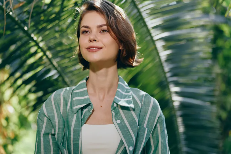 Portrait of a young woman outdoors under sunlight, wearing a striped green shirt, with lush tropical leaves in the background.