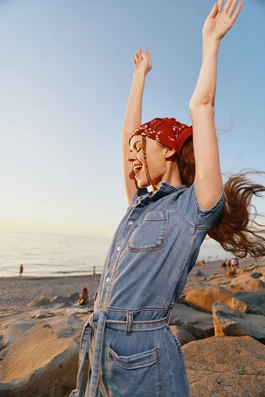 Lifestyle woman in modern boho-western denim street style joyfully raising arms by the beach during golden hour with film color effect enhancing warmth and natural light