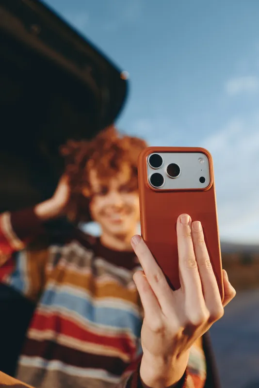 Woman lifestyle selfie outdoors, holding smartphone in one hand, phone closeup and screen facing camera, portrait of curly haired youth in striped shirt in sunlight blue sky