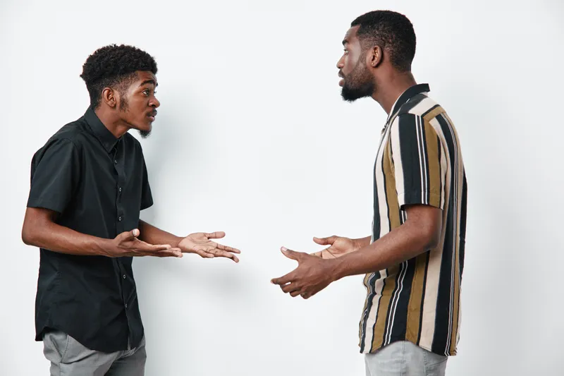 Two black men with dark skin having a serious conversation in casual clothes on white background. African ethnicity, communication, discussion, disagreement, standing indoors.