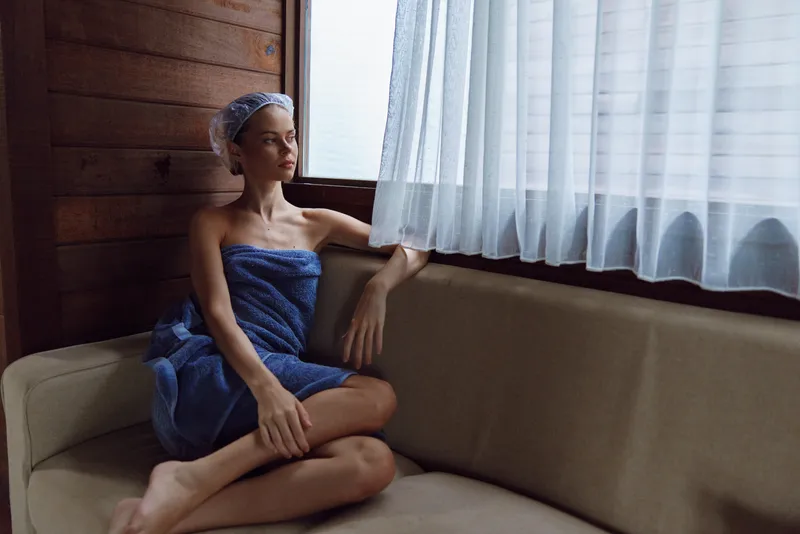 Woman in towel sitting thoughtfully on beige sofa near window with sheer curtain, natural light illuminating her calm face and relaxed posture indoors.