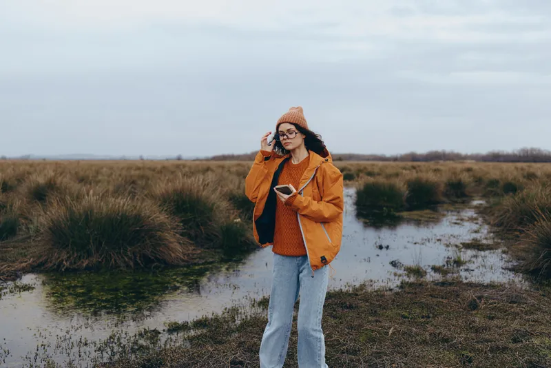 Woman lifestyle outdoors in an orange jacket and knit hat stands by a narrow river in a grassy field under cloudy sky, relaxed walking, casual travel moment in natural landscape.
