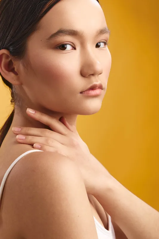 asian woman with smooth skin gazes softly at the camera in a close up portrait, warm yellow backdrop highlights natural beauty and serene confidence in a modern studio setting.