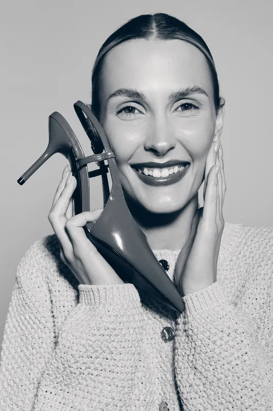 Smiling white woman with fair skin holds high heel shoe near face wearing knitted sweater studio portrait.
