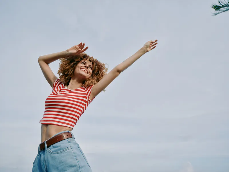 Young woman with curly hair dances outdoors, arms raised, wearing a red striped crop top and light denim jeans, smiling and carefree against a bright, clear sky, capturing energy and joy.