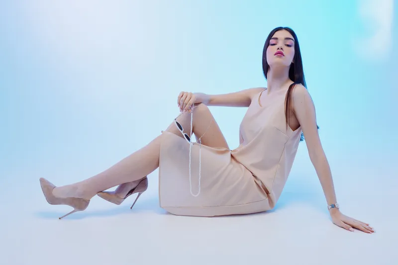 Elegant woman in a beige dress and high heels poses sitting on the floor against a light blue background. Concept of beauty, fashion, style, and modern elegance.