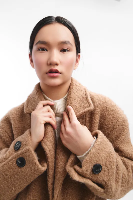 Asian woman wearing a camel coat and knit sweater stands in a clean studio, presenting a calm, confident look. The portrait emphasizes natural texture and warm fashion detail with soft lighting.