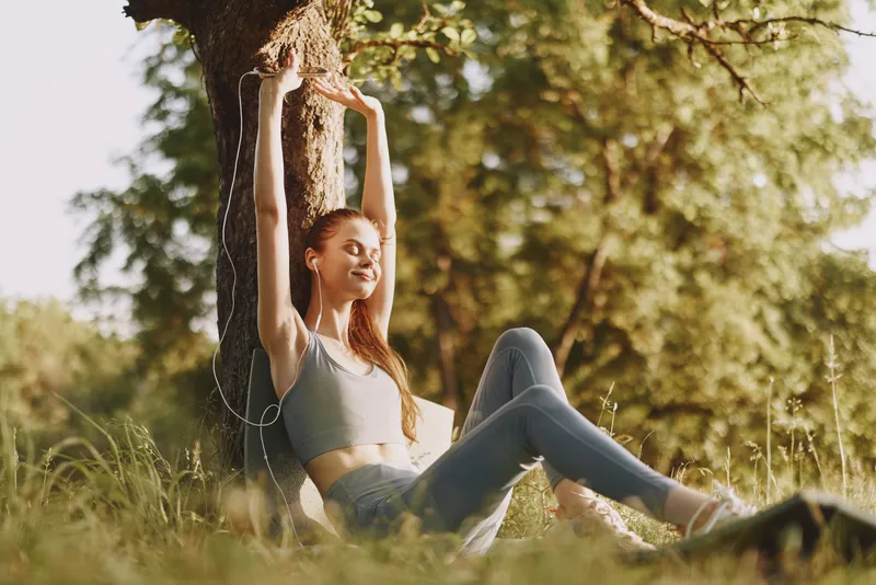 Young woman enjoying outdoor relaxation under a tree, wearing activewear and headphones, embodying tranquility and fitness in a natural, sunny environment