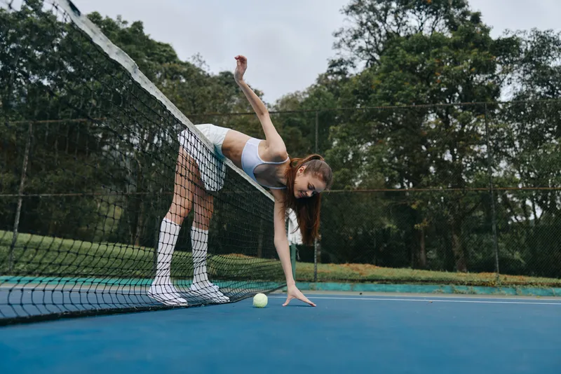 Young female athlete wearing sporty attire, bending over the tennis net, reaching for a ball, showcasing determination and focus in a lush outdoor setting