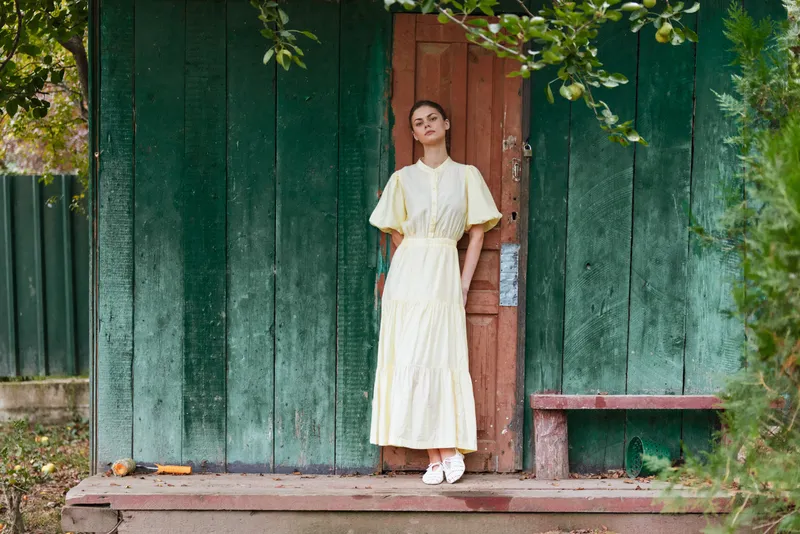 Woman in a pale yellow dress stands at a wooden doorway in a garden setting, creating a calm fashion moment outdoors with soft natural lighting in inspiration.