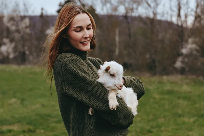 Joyful woman in a dark green sweater holds a fluffy white dog outdoors, sharing a tender moment in a grassy park, conveying warm companionship and gentle affection under daylight.