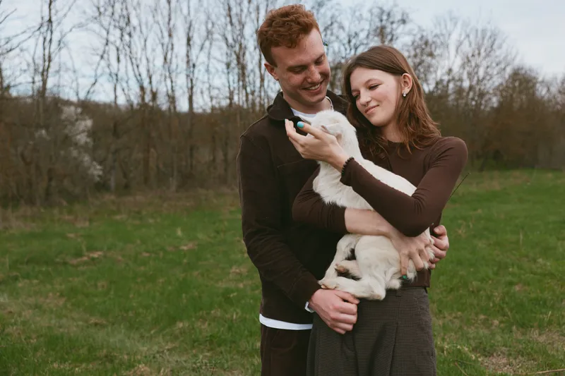 A smiling couple outdoors gently holds a young goat kid, sharing a tender moment in a sunlit field. A lifestyle scene that emphasizes companionship, animal care, and rural living, perfect for family