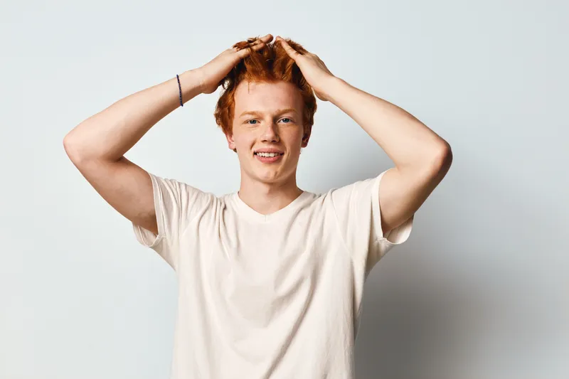 Young man with red hair smiling and hands on head wearing white t shirt against plain background. Happy male portrait with casual style and cheerful face expression in studio lighting.