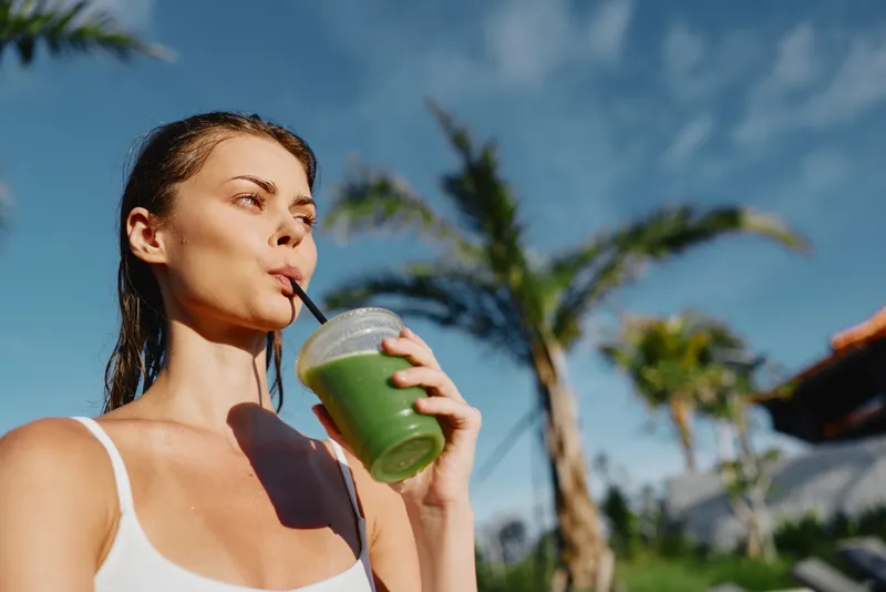 Healthy woman outdoors drinks a green smoothie, sunlight bright, wearing a white tank top, sipping through a straw, palms and blue sky behind, capturing a relaxed, vibrant summer lifestyle moment