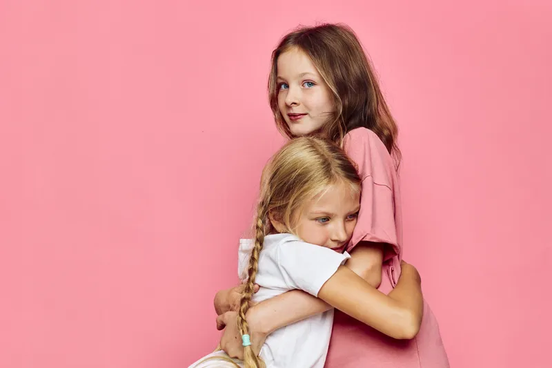 Two girls hugging each other on a pink background. One girl looks at the camera, the other with braided hair hugs tightly. Concept of family love, care, and childhood.
