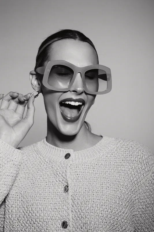 Stylish caucasian woman with fair skin wearing large sunglasses and knitted sweater laughs joyfully with open mouth on grey background.