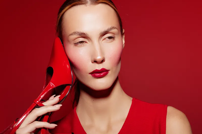 Woman with pale skin and red lipstick holds red high heel shoe near face on red background. Concept of fashion, style, beauty, and monochrome color theme.