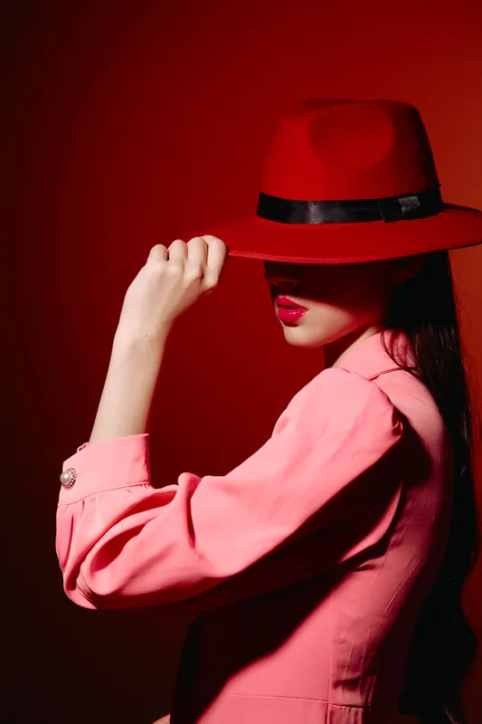 Stylish woman in red hat and pink blazer partially covering her face, creating mysterious and creative beauty fashion portrait with strong color contrast in studio light.