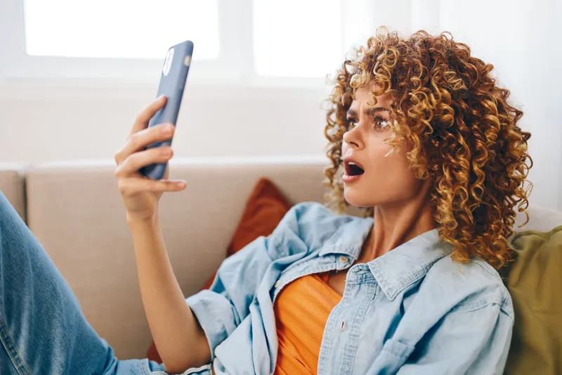 Улыбающаяся женщина держит смартфон, отдыхает на уютном диване дома, наслаждается онлайн-играми и общением в киберпространстве