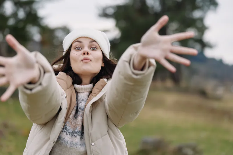 Woman reaching toward the camera outdoors in a warm jacket and beanie, playful and joyful in a wide natural field under soft light.