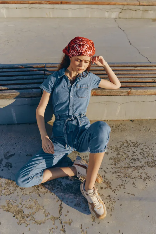 Lifestyle woman in modern boho-western denim street style wearing a red bandana, sitting casually outdoors under warm sunlight with film color tones.