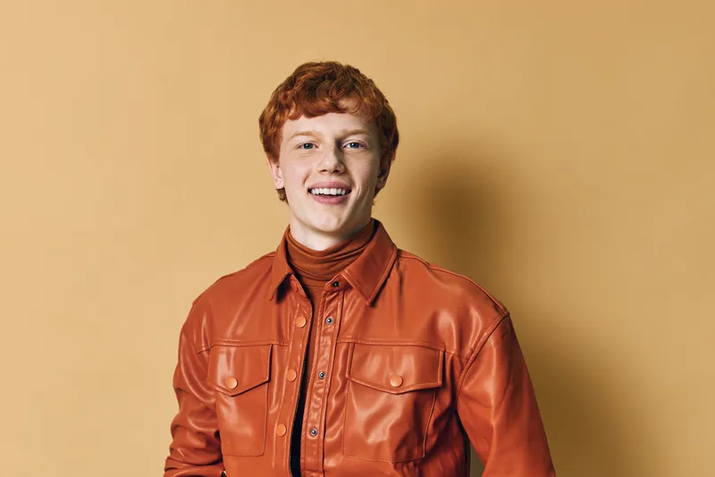 Young man with red hair wearing orange leather jacket and brown turtleneck smiling at camera against beige background in studio portrait.