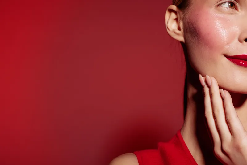 Close-up of a woman with red lipstick and red dress touching her face against a red background. Concept of beauty, makeup, fashion, and elegance with vibrant colors.