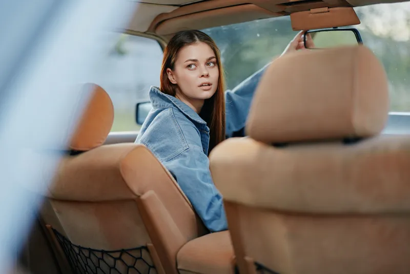Young woman in denim jacket looking back from the front seat of a car, conveying curiosity and contemplation during a road trip adventure.